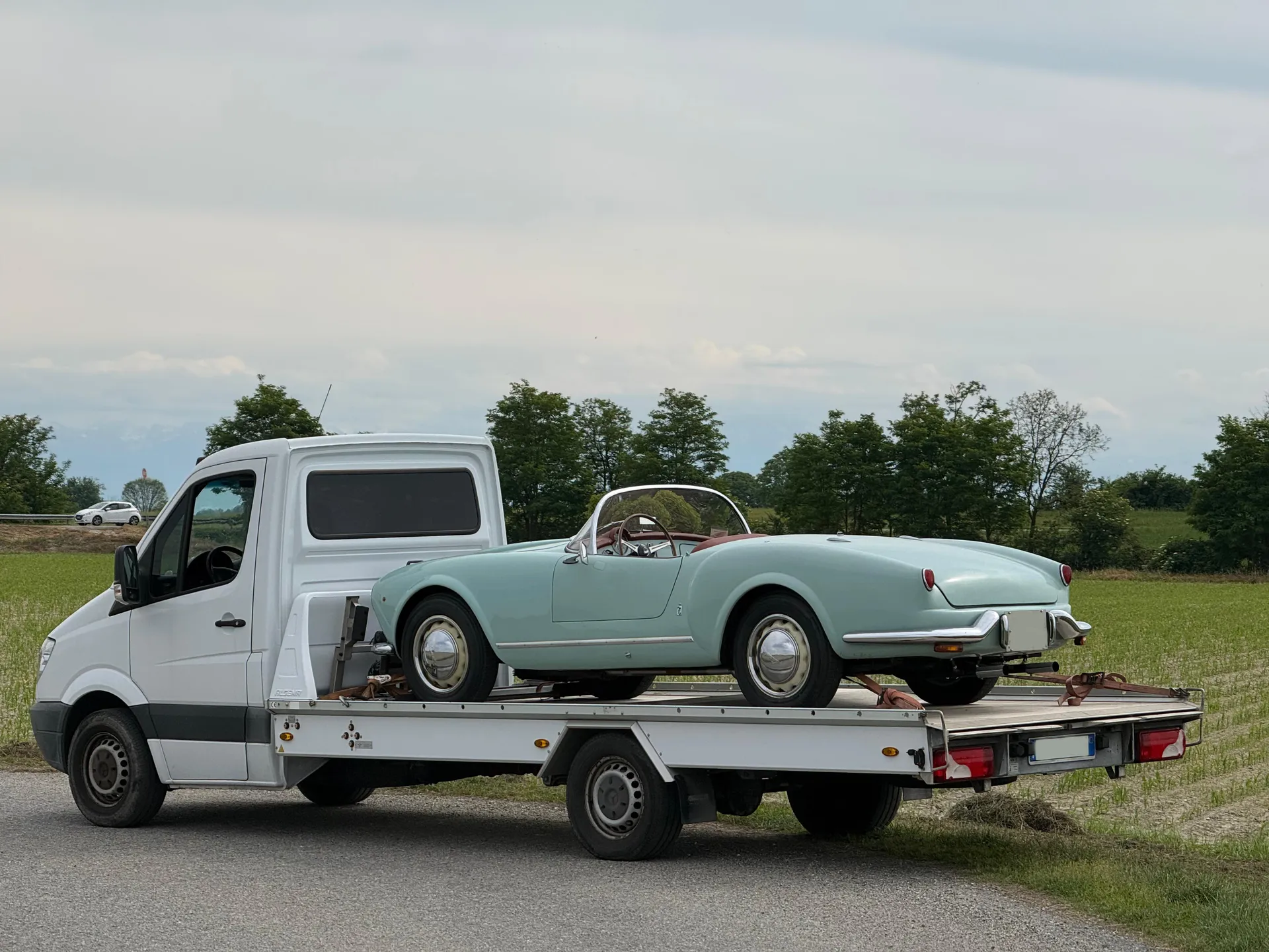 Lancia Aurelia B24 Spider America azzurro pastello vista dal retro su bisarca Mercedes Sprinter di Rubino Express lungo una strada di campagna